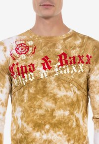 Langarmshirt aus braunem Batikstoff mit roten und cremfarbenen grafischen Texten und Emblemen auf der Brust und den Schultern.