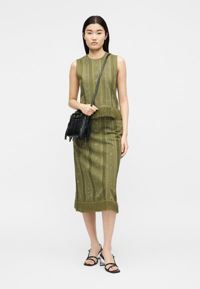 Γυναίκα που φορά αμάνικο ελαιώδες χρώμα φόρεμα με μοτίβα και λεπτομέρειες με φούντες, κρατώντας μικρή μαύρη τσάντα χιαστί και φοράει μαύρα σανδάλια με τακούνι.