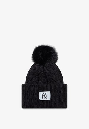 Cappello di lana nera lavorato a maglia con motivo a treccia, dotato di un pompon in ecopelliccia e un'etichetta bianca con logo sul davanti.