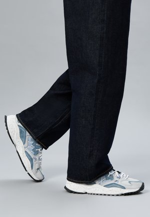 Person wearing white and gray speckled sneakers with blue accents and dark denim jeans, walking against a plain gray background.