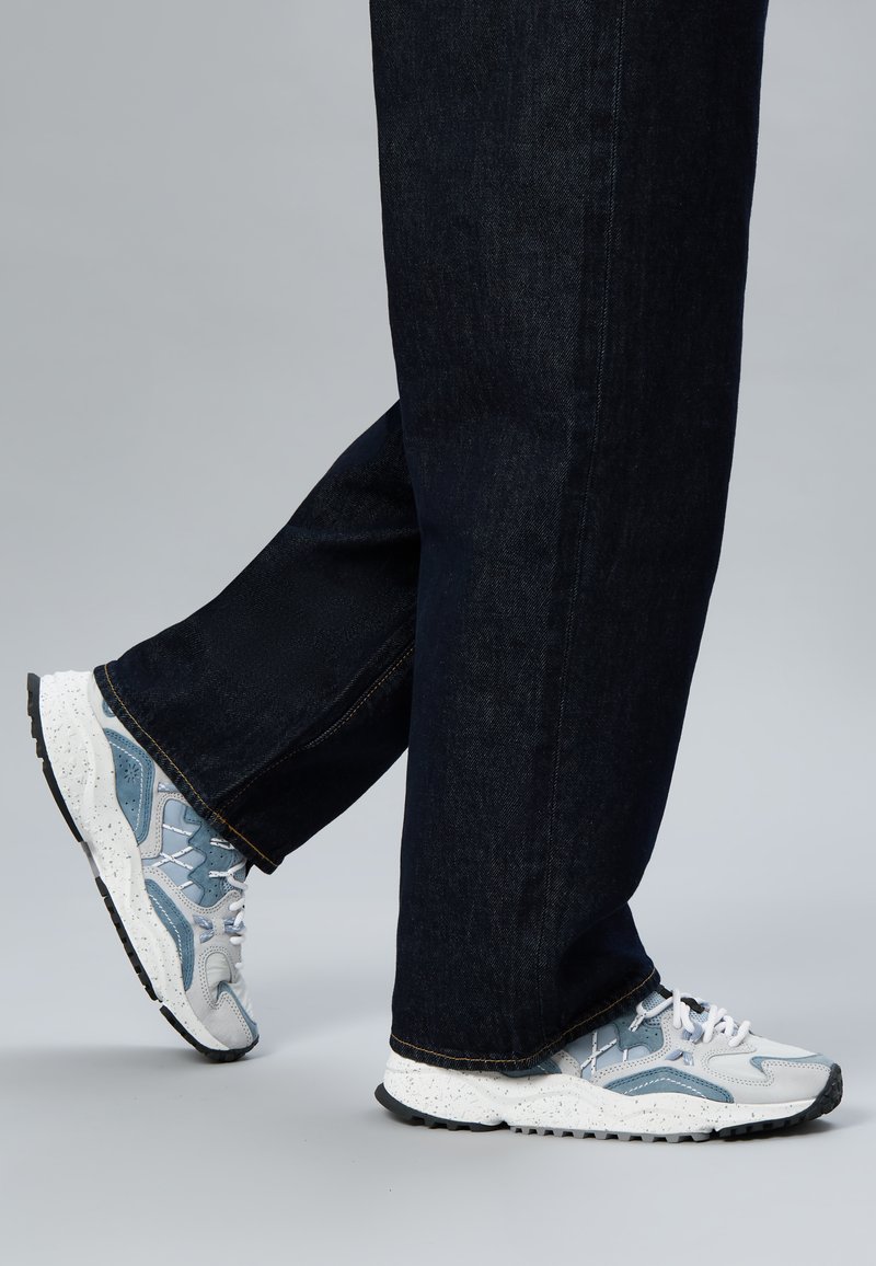 Persona que lleva zapatillas blancas y grises con moteado y detalles azules, y vaqueros oscuros, caminando sobre un fondo gris liso.