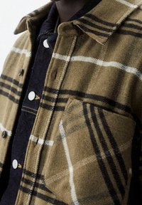 Camisa chaqueta de franela marrón de lana con un gran patrón de cuadros en blanco y negro, que presenta un bolsillo en el pecho y botones a presión plateados.
