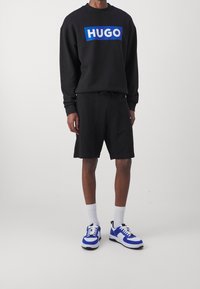 Felpa nera con logo blu "HUGO", abbinata a pantaloni corti neri. Calze corte bianche e sneaker atletici blu e bianchi completano l'outfit.