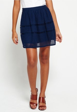 Falda azul marino hecha de tela perforada, con cintura elástica y diseño por capas. Combinada con sandalias de cuero marrón.