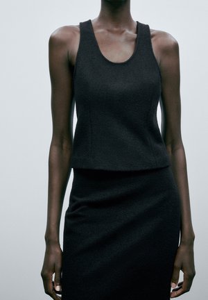 Femme portant un haut noir texturé sans manches et une jupe assortie sur un fond gris clair uni.