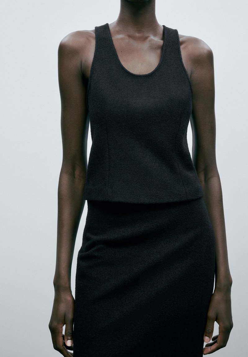 Femme portant un haut noir texturé sans manches et une jupe assortie sur un fond gris clair uni.