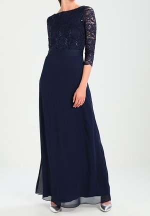 Vrouw draagt een marineblauwe avondjurk met kanten pailletten driekwart mouwen en zilveren hoge hakken, staand op een witte achtergrond.