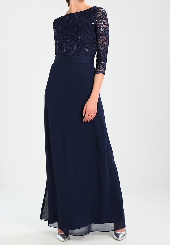 Femme portant une robe de soirée bleu marine avec manches trois-quarts en dentelle à sequins et chaussures à talons argentées, debout sur un fond blanc.