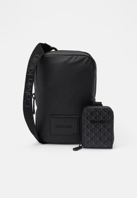 Geantă de umăr sintetică neagră cu curea ajustabilă, având un patch cu logo. Include un pouch cu fermoar asortat, cu un design pattern.