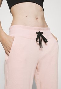 Kvinna iklädd ljusrosa sweatpants med svart dragsko och svart croptop, händerna i fickorna, överkroppen synlig mot en enkel bakgrund.