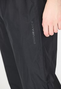 Sorte træningsbukser med en glat tekstur og diskret glans. Har et trykt "ENDURANCE" logo langs siden som brandingdetalje.