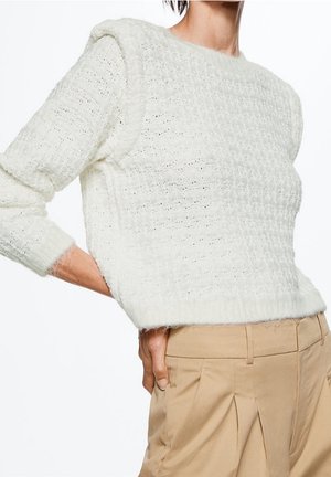 Vrouw draagt een textuurs witte gebreide trui met schoudervullingen en beige hoge taille plissébroek, hand rustend op de heup.