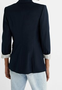 Blazer blu navy in tessuto liscio con una forma aderente, dettagli di polsini plissettati in grigio chiaro e uno spacco sul retro per facilitare i movimenti.