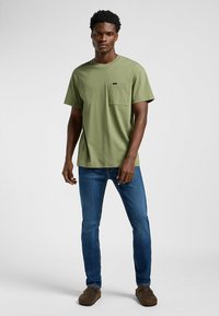 T-shirt in cotone verde oliva con tasca sul petto, abbinato a jeans di denim blu. Il modello indossa scarpe casual e si trova su uno sfondo neutro.