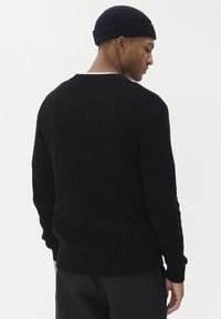 Man som bär en svart stickad tröja och mössa i matchande färg, vänd åt sidan med huvudet något nedåtvinklat framför en enfärgad vit bakgrund.