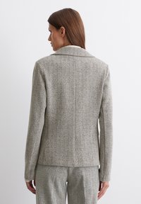 Grauer Blazer im Fischgrätmuster mit Reverskragen, taillierter Form und strukturierten Schultern. Glatte Textur ohne sichtbare Verschlüsse.