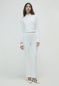 Donna con un maglione bianco a maniche lunghe, pantaloni bianchi dritti, bracciali d'oro e tacchi alti bianchi con lati trasparenti, in piedi su uno sfondo bianco.