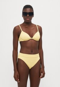 Gul ribbet bikinioverdel med tynne stropper og trekantet form, kombinert med høy midje ribbet bikinitruser. Teksturert stoff.
