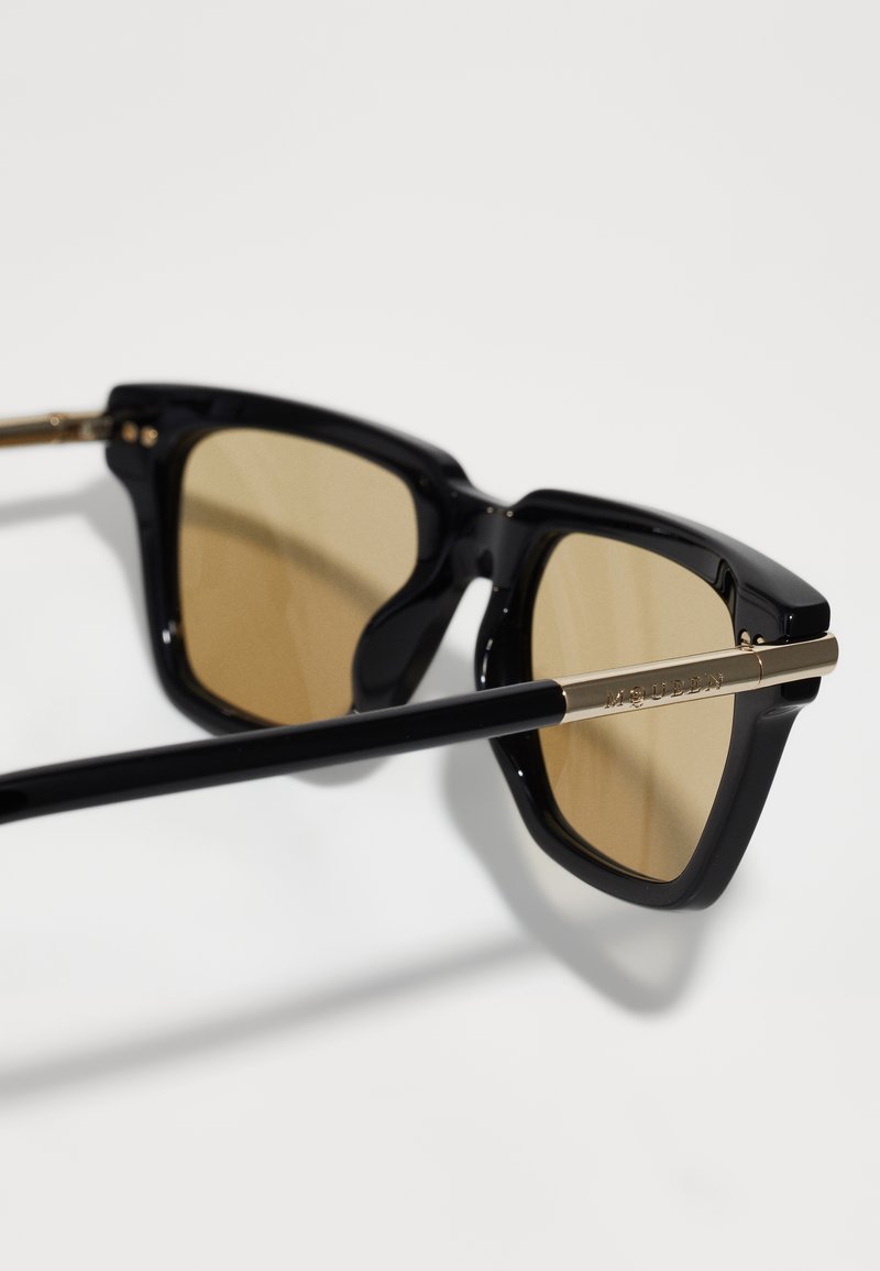 Schwarze Sonnenbrille mit einem markanten, eckigen Rahmen, braun getönten Gläsern und goldmetallischen Akzenten an den Schläfen und Bügeln.