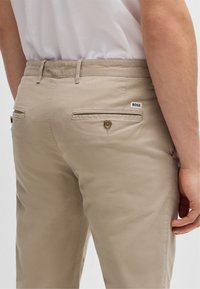 Beige pamut nadrág, sima textúrával, két hátsó zsebbel, gombos záródással, és egy kis fekete-fehér "BOSS" címkével.