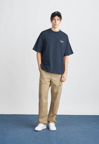 T-shirt oversize bleu marine avec un petit motif, pantalon cargo beige avec poches, et baskets blanches. Le modèle porte une casquette noire et se tient sur un tapis bleu.