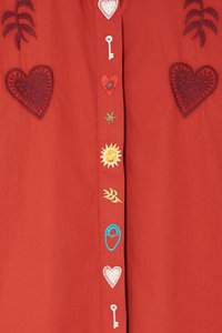 Punainen paita, jossa on brodeerattuja sydämiä, avain, aurinko ja lehtimotiiveja keskeisen nappilistansa ympärillä. Koristeellisia tekstuureja ja kuvioita on läsnä.