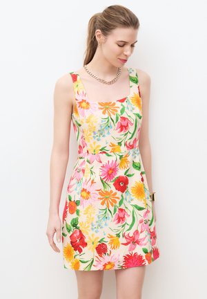 Kobieta ubrana w sukienkę bez rękawów z kwadratowym dekoltem w żywy, wielokolorowy kwiatowy wzór, złoty łańcuszek i bransoletka na tle białego tła.