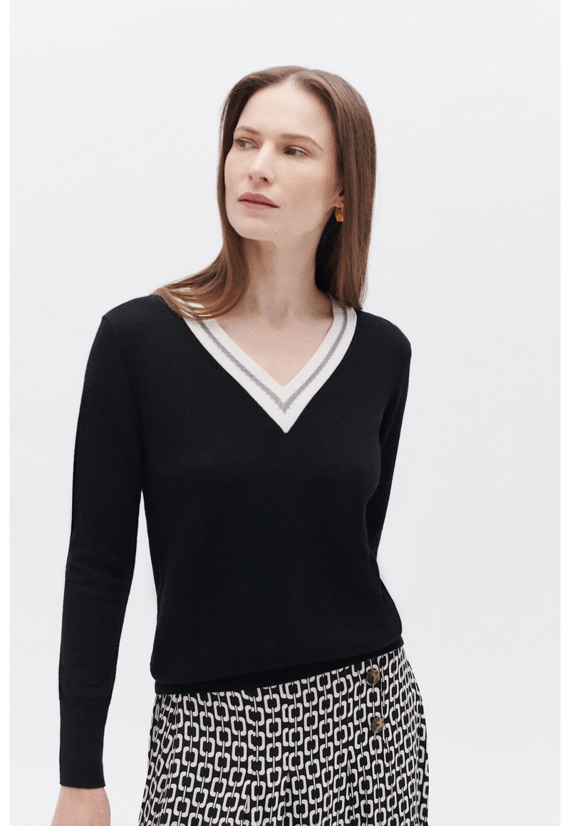 Maglione nero a V con bordi bianchi e grigi a contrasto, caratterizzato da maniche lunghe e una forma aderente, abbinato a pantaloni con fantasia.