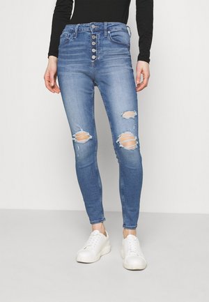 Jeans en denim bleu taille haute avec une fermeture à cinq boutons, des genoux déchirés et un design slim, associés à des baskets blanches.