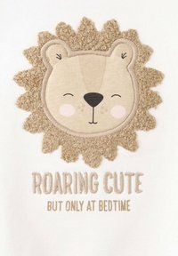 Un tessuto bianco presenta un design di testa di leone con pelo beige texturizzato, guance arrossate e testo ricamato: "RUGGENTE E CARINO MA SOLO A ORARIO DI NANDA".
