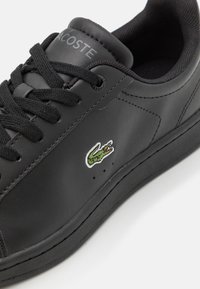 Fekete bőr sneaker, fűzős záródással, textúrált felülettel, gumi talppal és zöld krokodil logóval az oldalon. Látható varrási részletek.