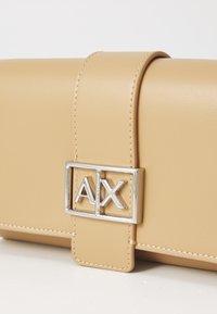 Beige kunstlærveske med en skinnende sølv rektangulær logo-lås med "AX." Glatt tekstur og quiltede detaljer langs kanten.