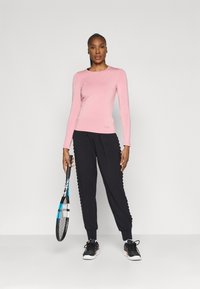 Light pink fitted long-sleeve top with scoop neck, paired with black sweatpants featuring scalloped accents, holding a tennis racket.