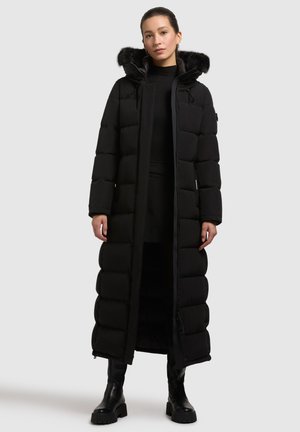 Langer schwarzer Daunenmantel mit einer mit Fell gefütterten Kapuze, mit gestepptem Design, einem Reißverschluss vorne und seitlichen Taschen. Getragen über einem schwarzen Outfit.