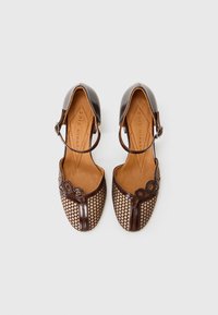 Scarpe in pelle marrone con design perforato, cinturino regolabile alla caviglia e dettagli circolari sulla parte frontale. Interno in pelle naturale.