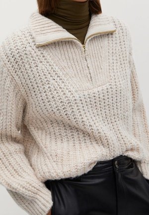 Pull en tricot épais écru avec fermeture éclair quart de tour et tirette anneau doré sur un col roulé marron foncé, associé à un pantalon en cuir noir taille haute.