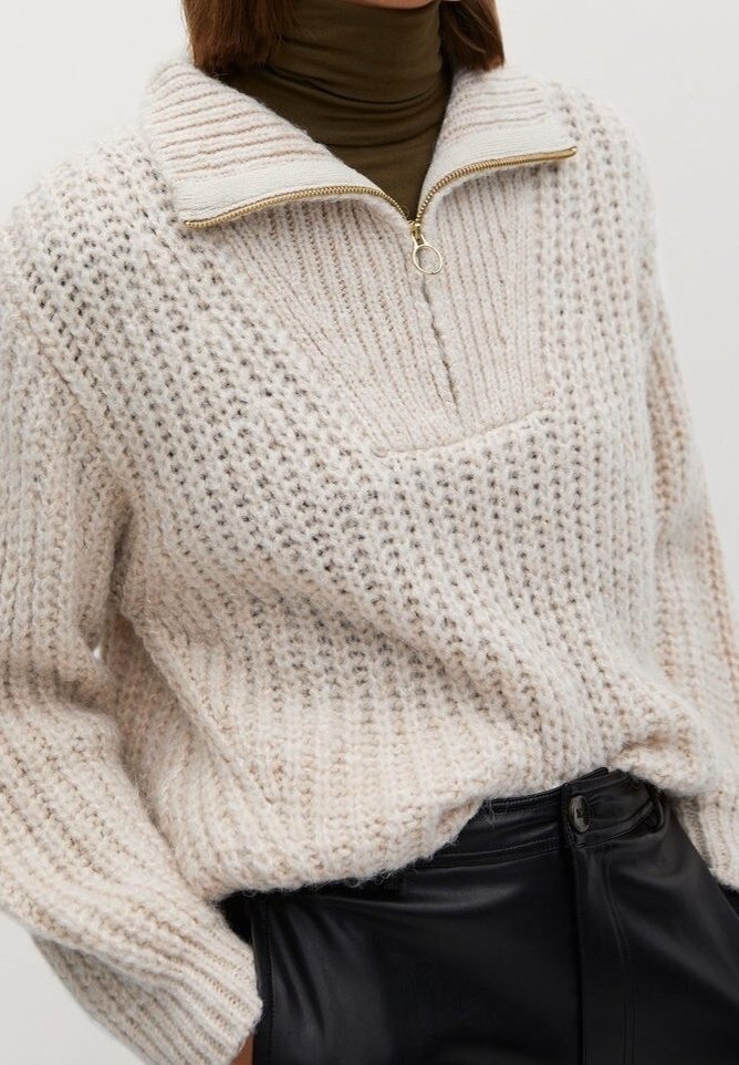 Pull en tricot épais écru avec fermeture éclair quart de tour et tirette anneau doré sur un col roulé marron foncé, associé à un pantalon en cuir noir taille haute.