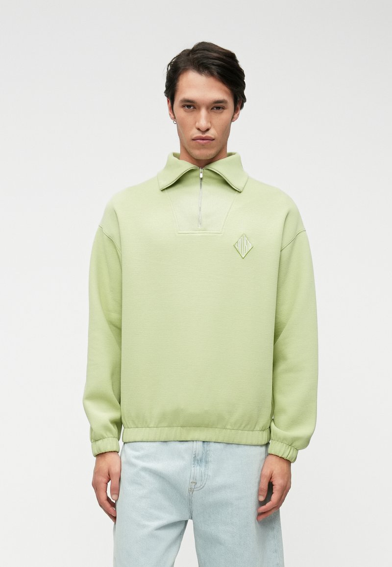 Ανοιχτό πράσινο φούτερ με φερμουάρ και ψηλό κολάρο. Διαθέτει λογότυπο μονόκερου στο στήθος και ραμμένα μανίκια, συνδυασμένο με ανοιχτό τζιν παντελόνι.