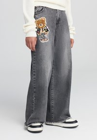 Jeans in denim grigio a gamba larga con orlo sfrangiato, caratterizzati da una patch in tessuto a forma di orso sulla coscia sinistra e abbinati a sneaker nere e bianche.