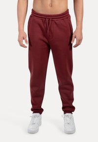 Maroons joggerbukser med elastisk talje, sidelommer og manchetter ved anklerne, parret med hvide sneakers. Glat stofstruktur.