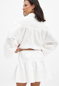 Blouse en lin blanc avec de larges manches cloche et un dos froncé. Associée à une jupe évasée assortie avec une taille plissée.