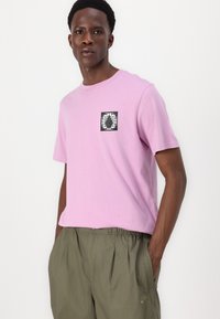 Lyserød bomulds-t-shirt med korte ærmer og et kvadratisk sort-hvidt ananasprint på brystet, sammen med løstsiddende olivengrønne shorts.