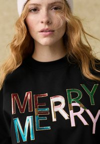 Fekete pulóver, amelyen színes flitterekből készült betűk formálják a "MERRY" szót különböző betűtípusokban. Az anyag sima textúrájú, és kerek nyakkivágása van.