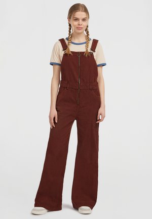 Giovane donna con capelli intrecciati, che indossa una t-shirt beige e delle salopette di velluto a coste bordeaux con zip sul davanti, abbinate a scarpe da ginnastica bianche, in piedi contro uno sfondo bianco.