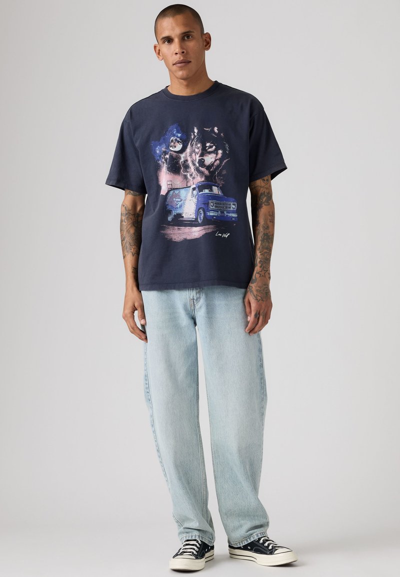 Uomo con tatuaggi che indossa una maglietta grafica scura con stampa di lupo e furgone, jeans azzurri e scarpe da ginnastica nere, in piedi davanti a uno sfondo semplice.