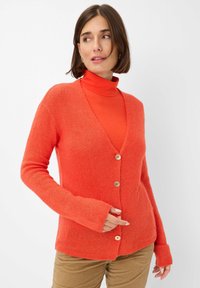 Strickjacke in Orange mit V-Ausschnitt, vorderen Knöpfen und langen �Ärmeln mit Daumenlöchern. Kombiniert mit einem passenden Rollkragenpullover darunter.