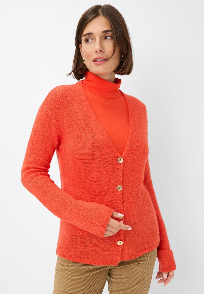 Strickjacke in Orange mit V-Ausschnitt, vorderen Knöpfen und langen Ärmeln mit Daumenlöchern. Kombiniert mit einem passenden Rollkragenpullover darunter.