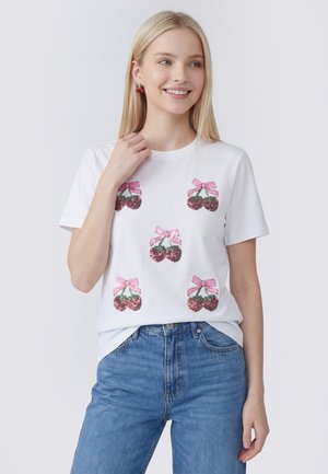 Jonge vrouw met steil blond haar draagt een wit T-shirt versierd met roze pailletten kersontwerpen en een blauwe spijkerbroek, licht glimlachend naar de zijkant.