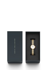 Montre en métal doré avec un cadran blanc rond, enfermée dans une boîte cadeau noire avec des lettres dorées et un intérieur rembourré.