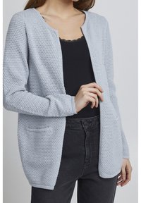 Light blue knitted cardigan with a textured pattern, open front, and two pockets, layered over a black tank top and dark jeans.